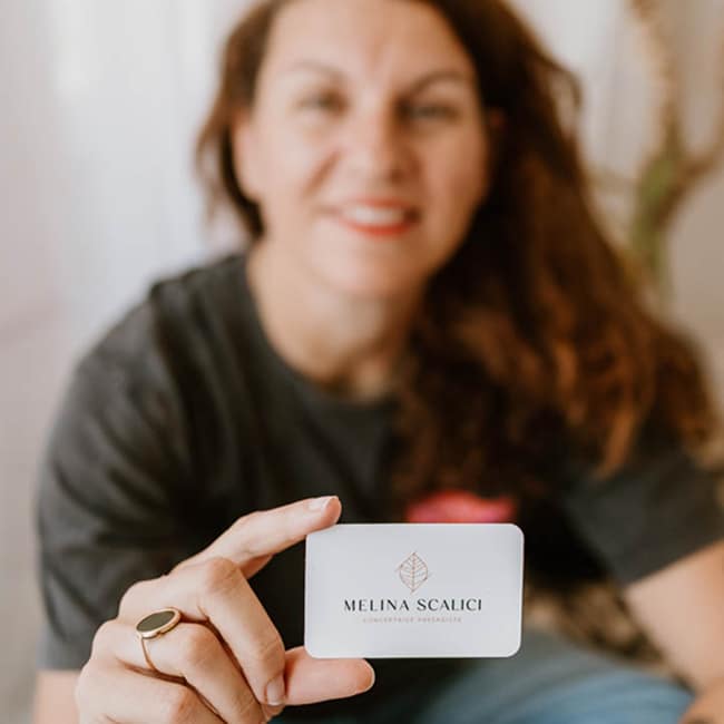 Melina Scalici, Conceptrice Paysagiste, présente sa carte Femme souriante présentant une carte de visite blanche et translucide avec le logo d'une feuille et le texte "MELINA SCALICI CONCEPTRICE PAYSAAGISTE".